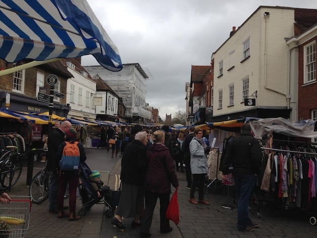 A wide shot of a bustling Elizabethan market, filled with colorful stalls, vendors hawking their wares, and crowds of people in period costumes. The scene is lively and chaotic, capturing the energy of the era.